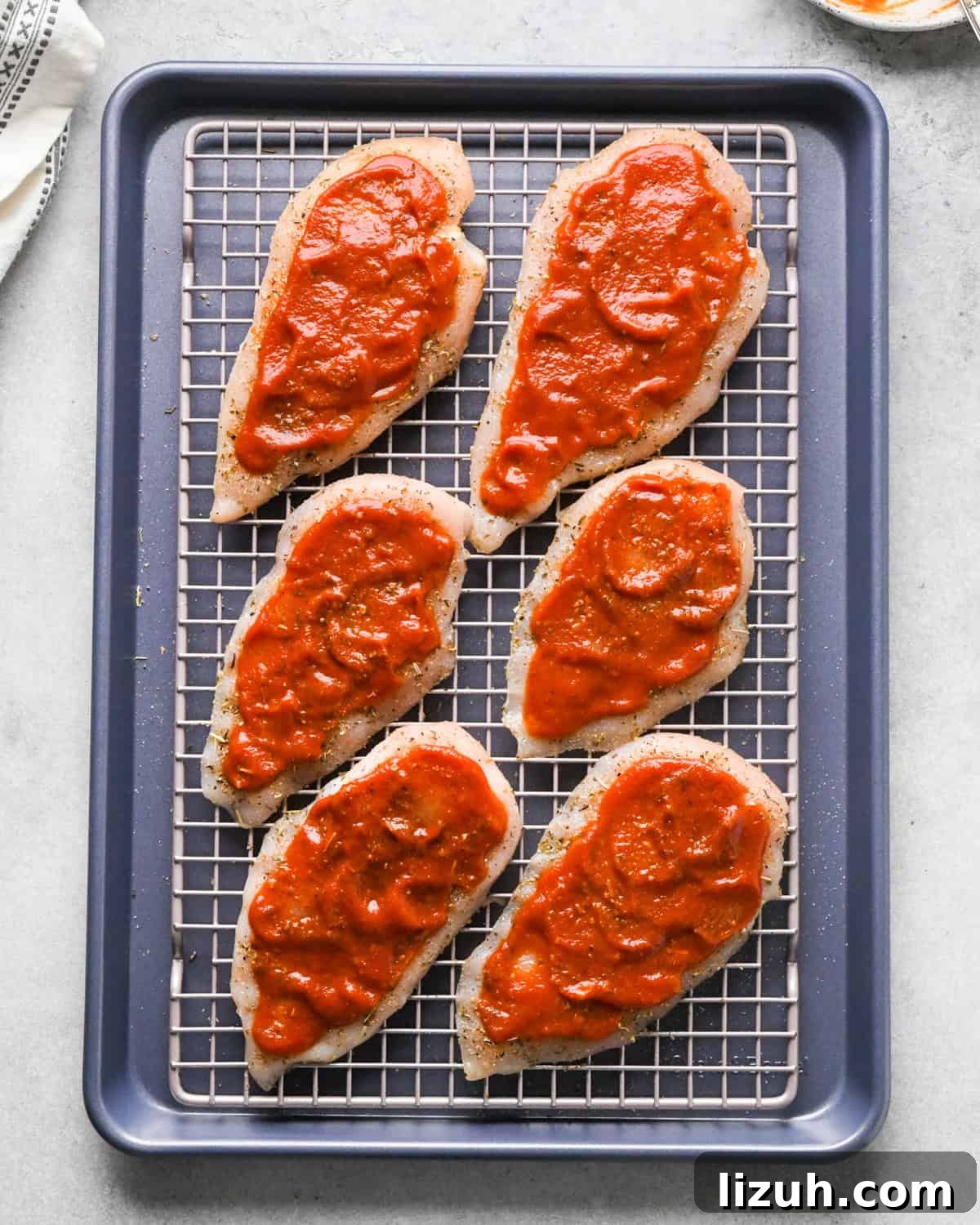 Making pizza chicken: overhead photo of chicken breasts sprinkled with spices and topped with pizza sauce. The chicken is sitting on a cooling rack on top of a baking sheet.