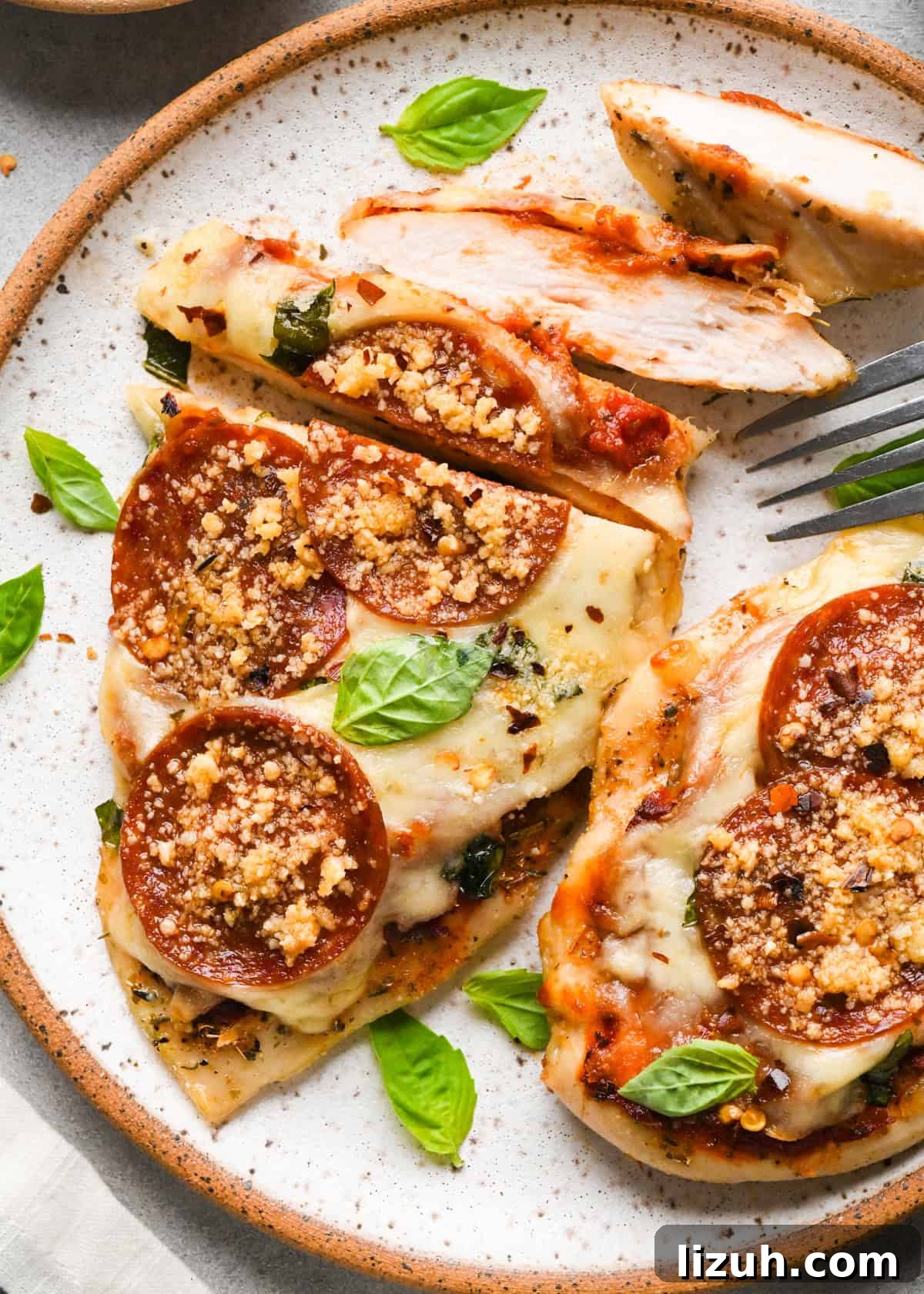 Plate of baked pizza chicken topped with melted mozzarella, pepperoni slices, Parmesan cheese, and fresh basil leaves, with a fork cutting into a piece.