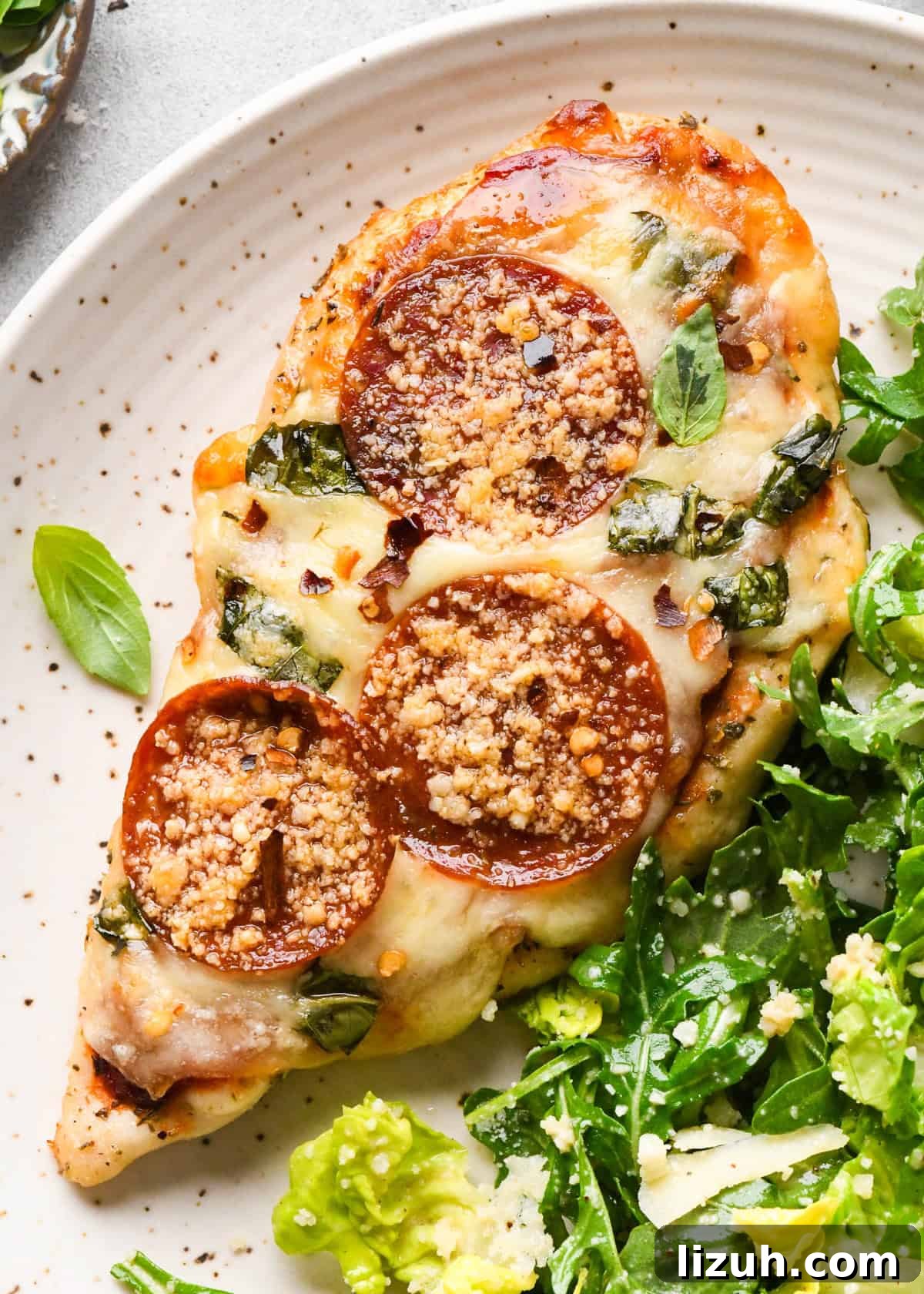 Close-up of baked pizza chicken topped with melted mozzarella, pepperoni slices, chopped basil, and grated Parmesan, served with a side of fresh green salad.
