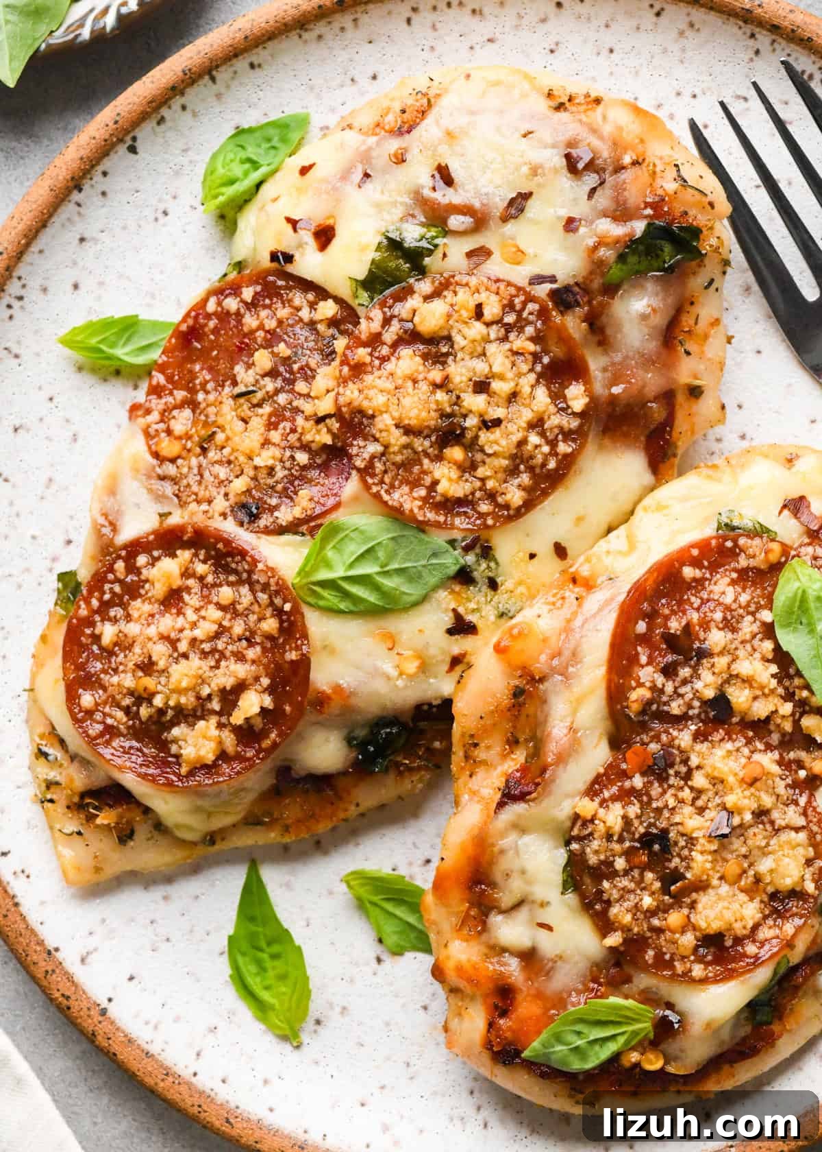 Plate of baked pizza chicken topped with melted mozzarella, pepperoni slices, Parmesan cheese, and fresh basil leaves, with a fork sitting on the plate.