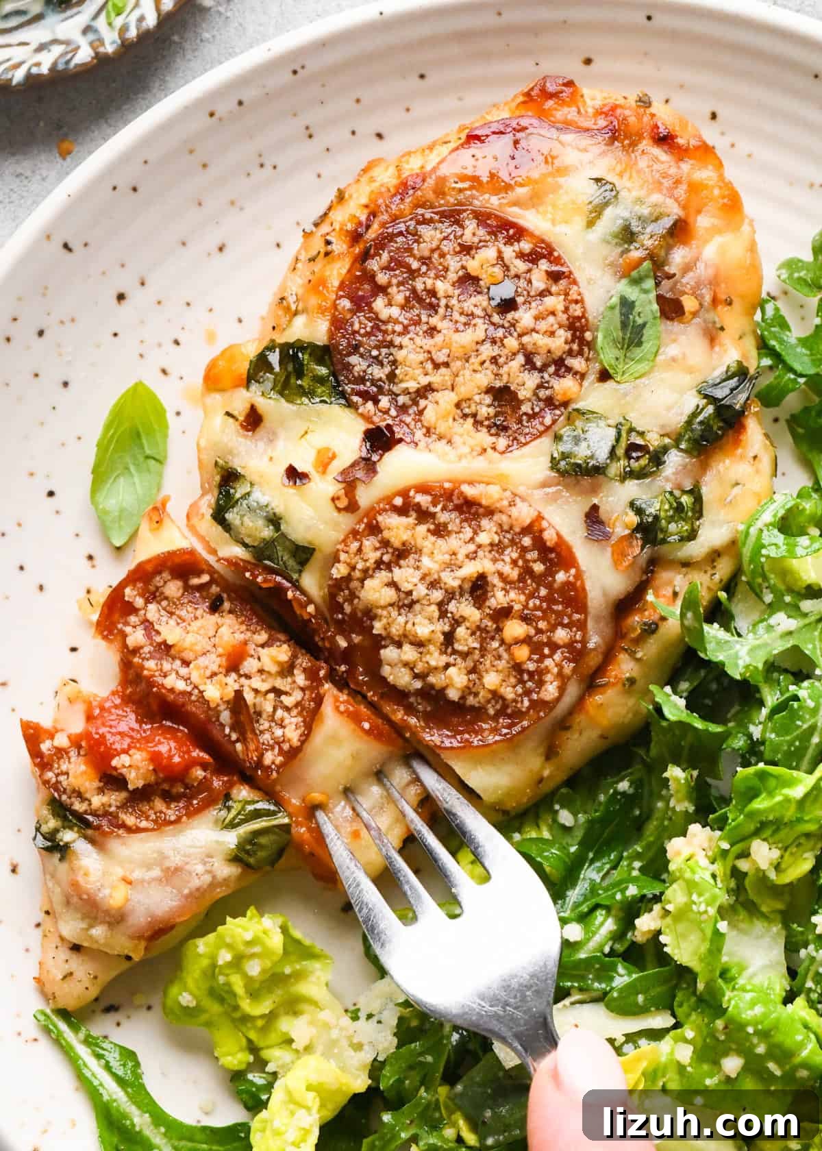 Close-up of baked pizza chicken topped with melted mozzarella, pepperoni slices, chopped basil, and grated Parmesan, served with a side of fresh green salad with a fork.
