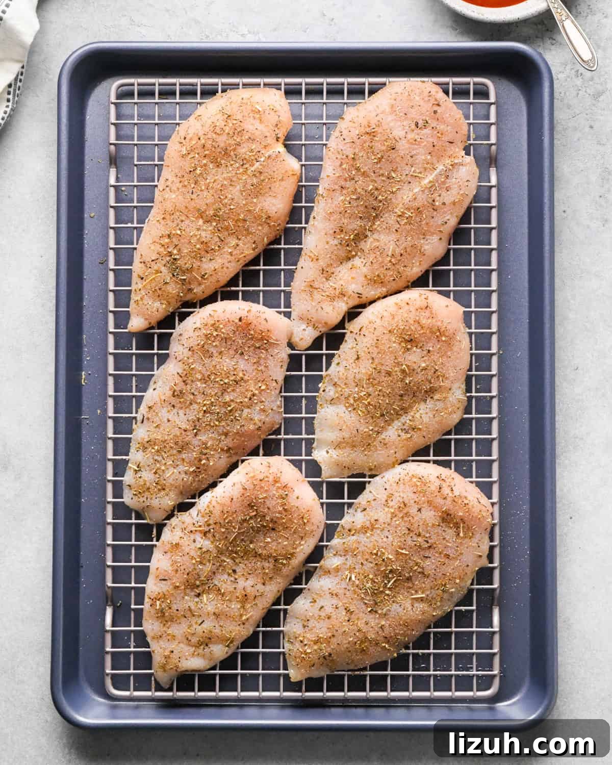 Making pizza chicken: overhead photo of chicken breasts sprinkled with spices and placed on a cooling rack on top of a baking sheet.